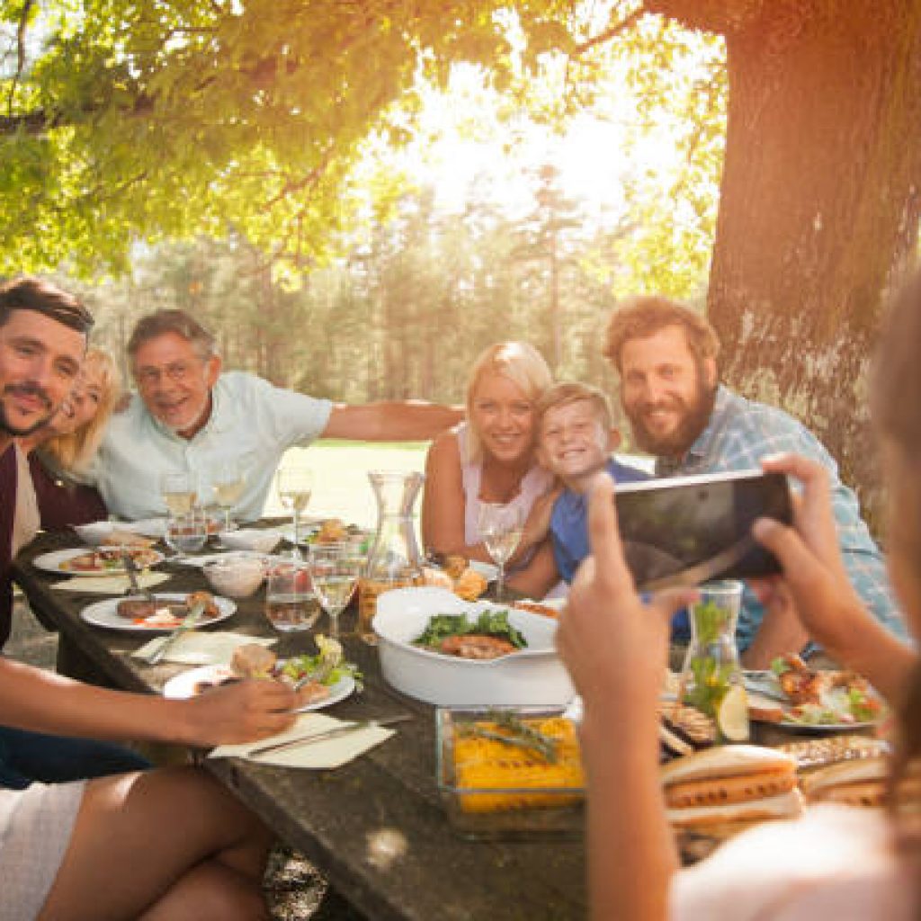 Grandfather taking a photo of his whole family on his smart phone at outdoor barbecue.